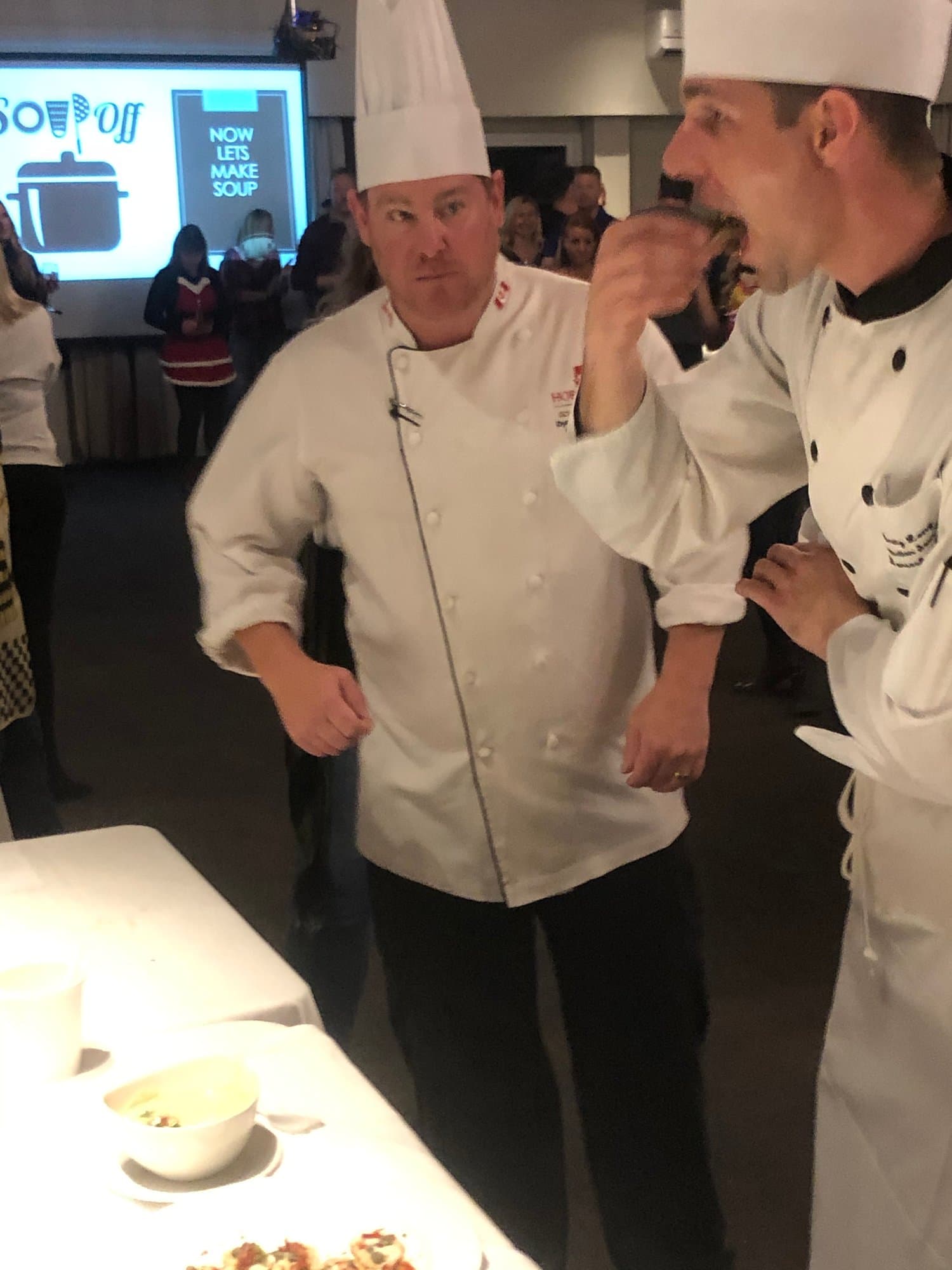 Two chefs in white jackets and tall hats at the judging table, one taking a bite while the other looks on