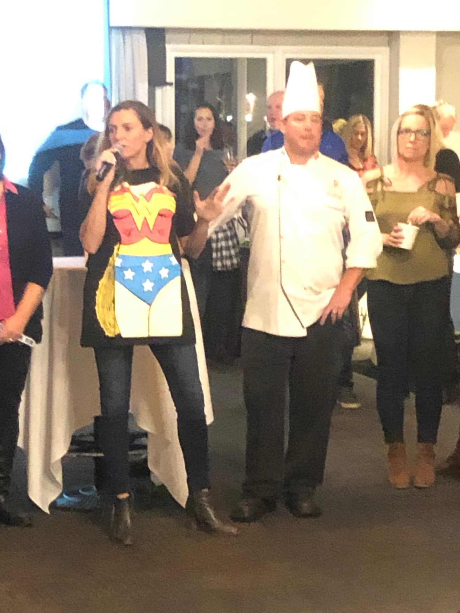 A speaker in a Wonder Woman apron talking into a microphone next to a chef as the crowd looks on
