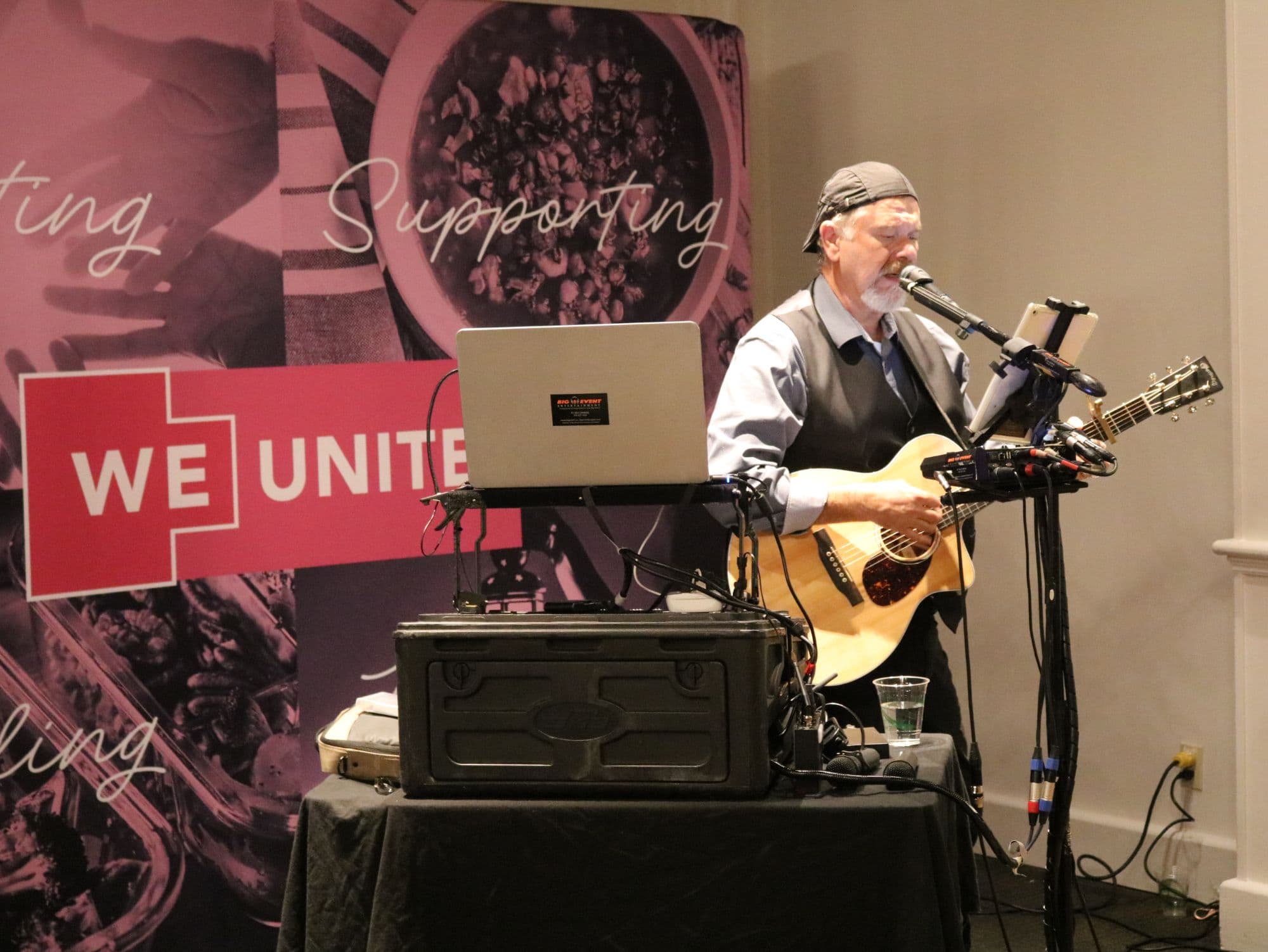 A musician singing and playing acoustic guitar in front of the WE Unite banner