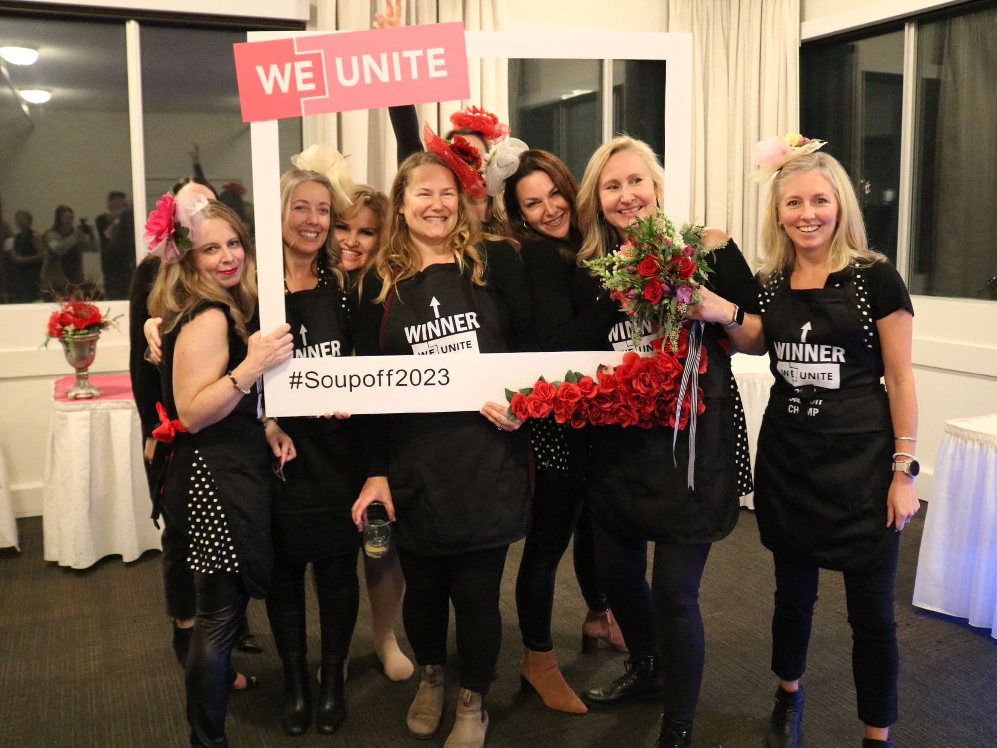 The winning team in matching Winner aprons and fascinators, holding roses in the WE Unite #Soupoff2023 photo frame